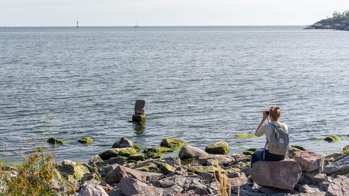 Person sitting on a rock by a shore, looking through binoculars at a rock installation at a distance in shallow water