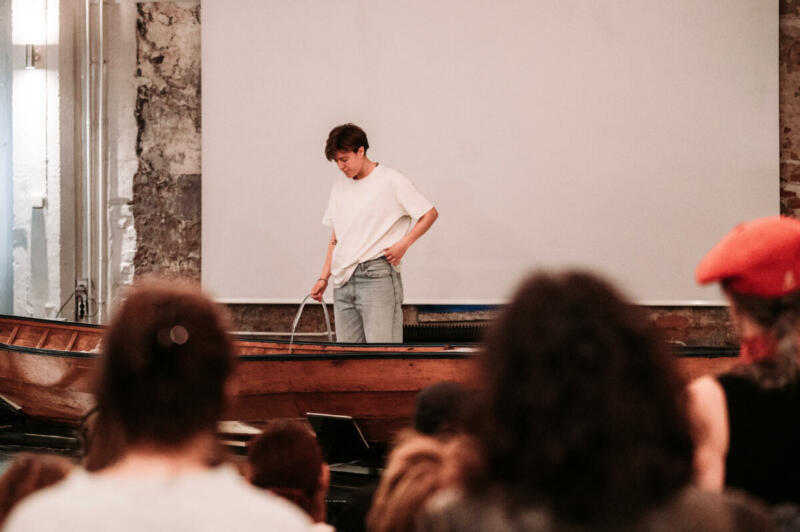 Stefanía is standing behind a wooden boat, filling the boat with water through a hose. People in the audience are watching.