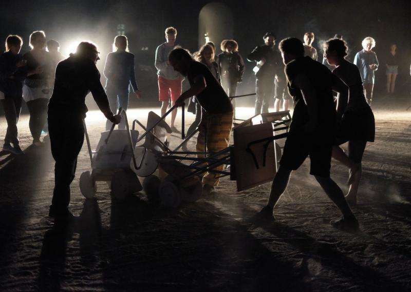 Four people are pushing around trolleys and bumbing into each other. In the background, a line of people are watching. It's dark and, there is only silhuettes of people.